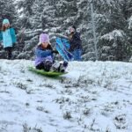 A person sledding down a snowy hill with others standing nearby in colorful winter clothing.