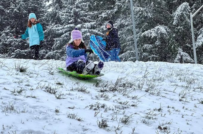 A person sledding down a snowy hill with others standing nearby in colorful winter clothing.