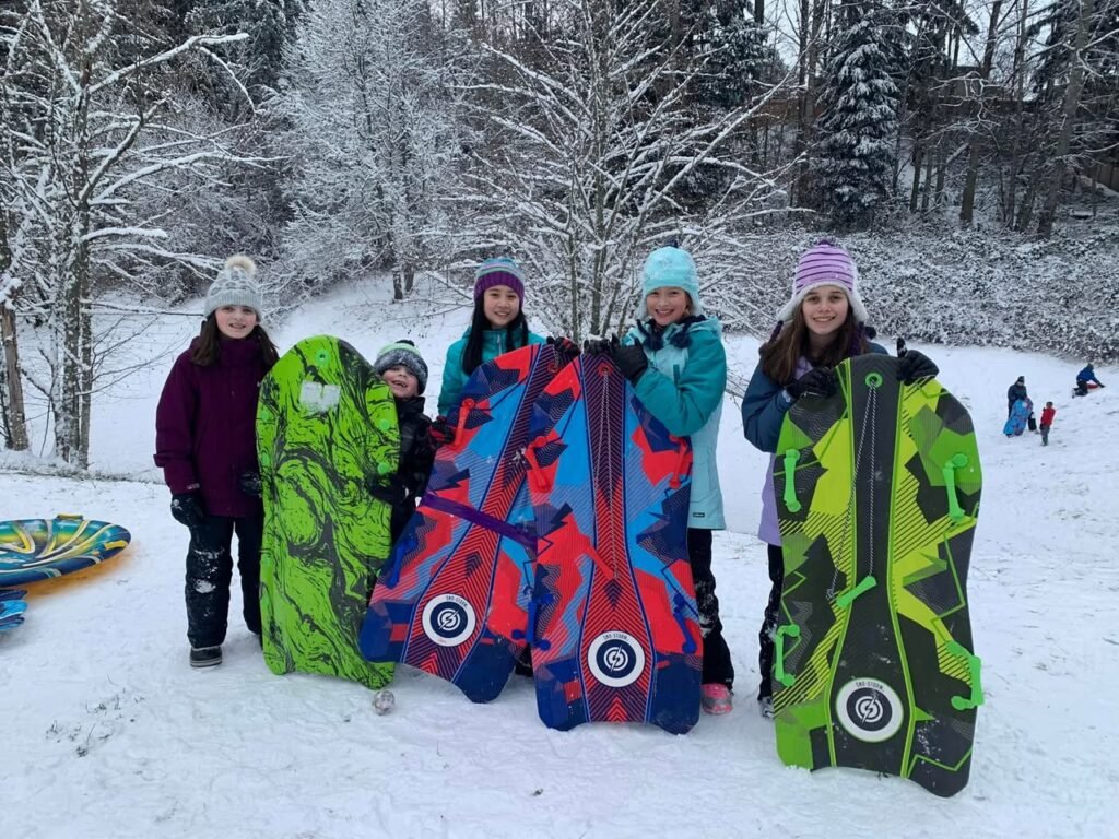 A group of snowsuit-wearing snowmobile riders holding colorful sleds in a frozen forest. The bright sleds stand out against the snow-covered landscape.