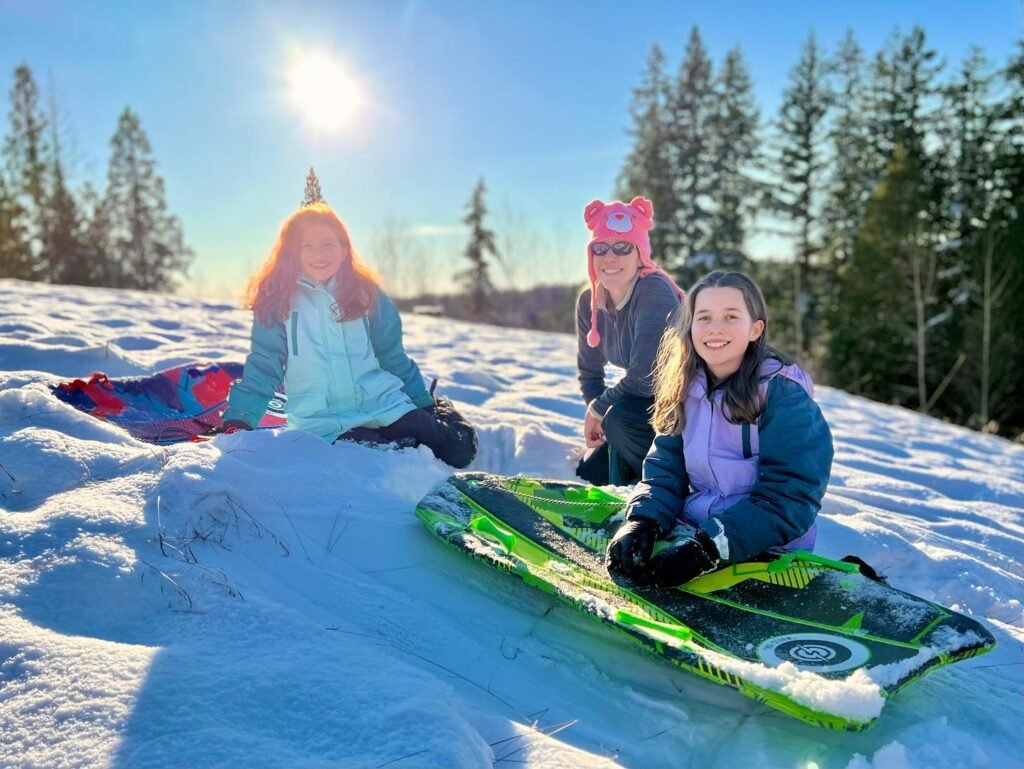 Three individuals pose with green sleds on a snow-covered hill under a bright sun. Tall evergreen trees line the background on a crisp winter day.