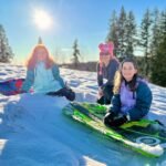 Let It Snow Three individuals pose with green sleds on a snow-covered hill under a bright sun. Tall evergreen trees line the background on a crisp winter day.