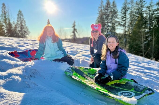 Three individuals pose with green sleds on a snow-covered hill under a bright sun. Tall evergreen trees line the background on a crisp winter day.
