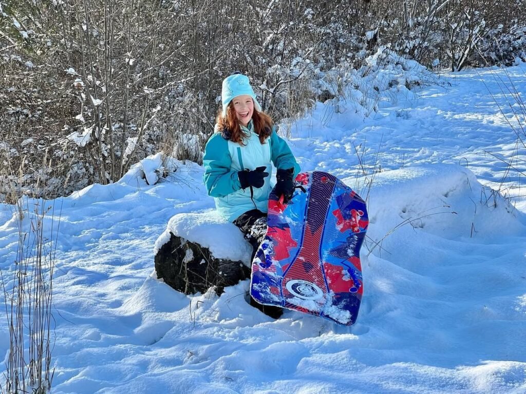 A child in a teal winter jacket sits on a snowy hill holding a colorful sled and snowshoes.