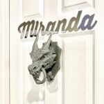 A close-up of a white door featuring the name Miranda in cursive script and a silver dragon-head door knocker. The decorative elements add a whimsical touch to the exterior.