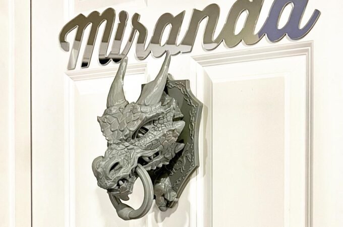 A close-up of a white door featuring the name Miranda in cursive script and a silver dragon-head door knocker. The decorative elements add a whimsical touch to the exterior.