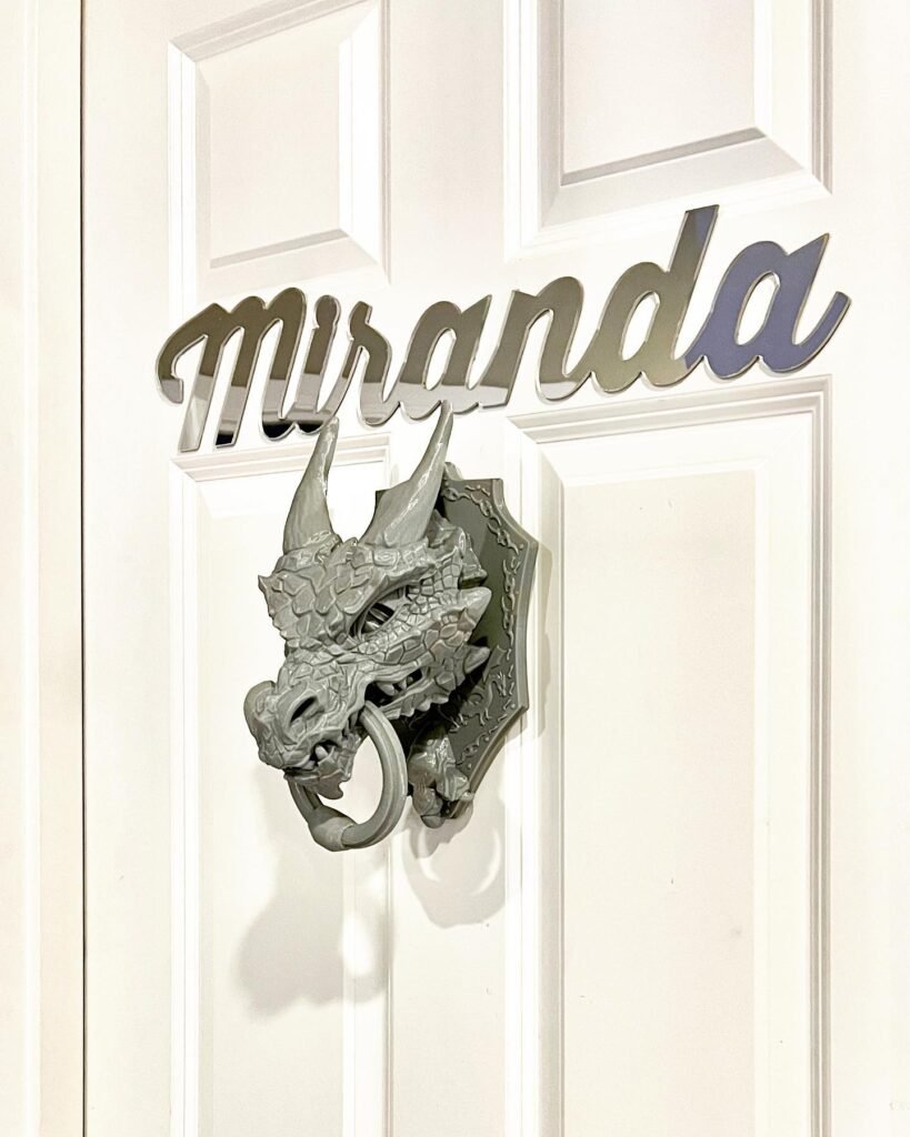A close-up of a white door featuring the name Miranda in cursive script and a silver dragon-head door knocker. The decorative elements add a whimsical touch to the exterior.