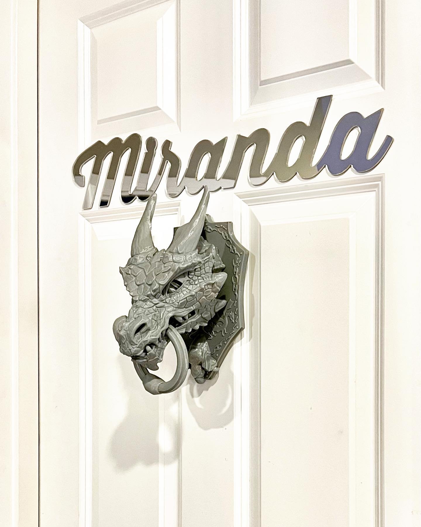 A close-up of a white door featuring the name Miranda in cursive script and a silver dragon-head door knocker. The decorative elements add a whimsical touch to the exterior.