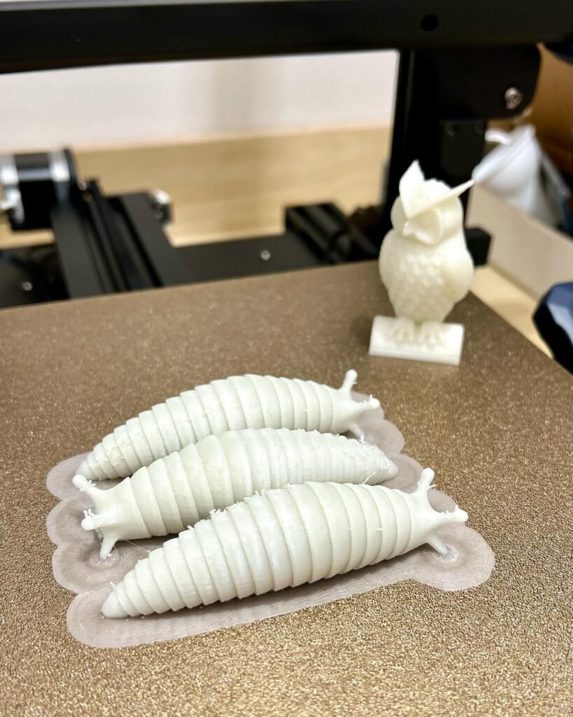 Close-up of three white 3D-printed chrysalises and a small owl figure, freshly printed on a workshop workbench.