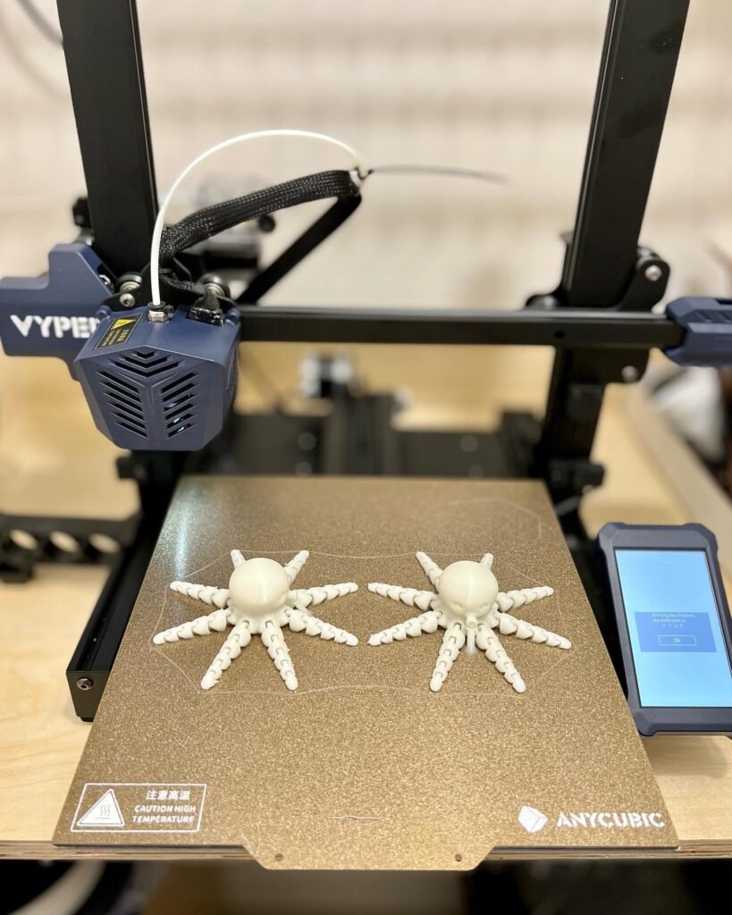 A 3D printed white spider ornament resting on a 3D printer's cardboard build plate. The Vyper extruder is visible in the background, positioned above the print surface.