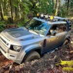 Fastpitch A silver Land Rover Defender 110 navigating a muddy trail in a dense forest.
