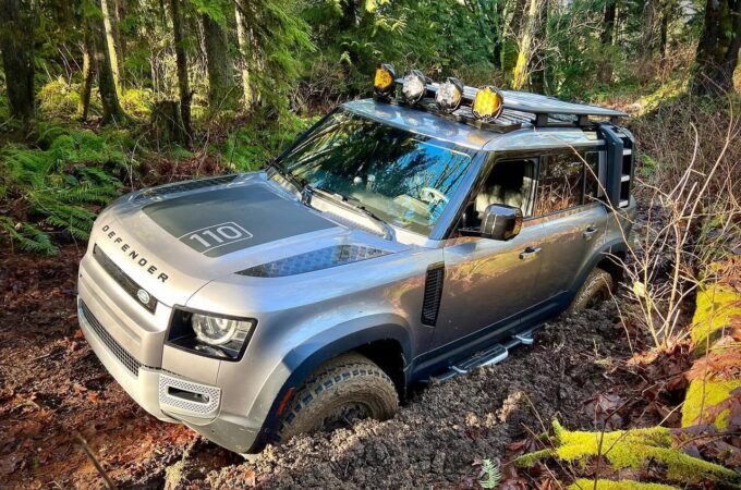 A silver Land Rover Defender 110 navigating a muddy trail in a dense forest.