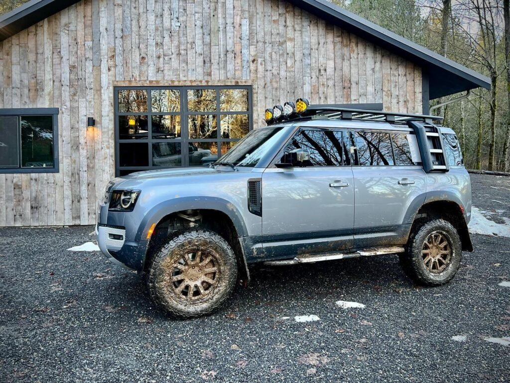 A muddy blue SUV parked in front of a rustic wooden building. The vehicle features off-road tires and roof-mounted lights.