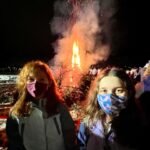 Two children wearing face masks stand in front of a large fire at night.