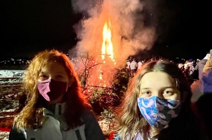 Two children wearing face masks stand in front of a large fire at night.
