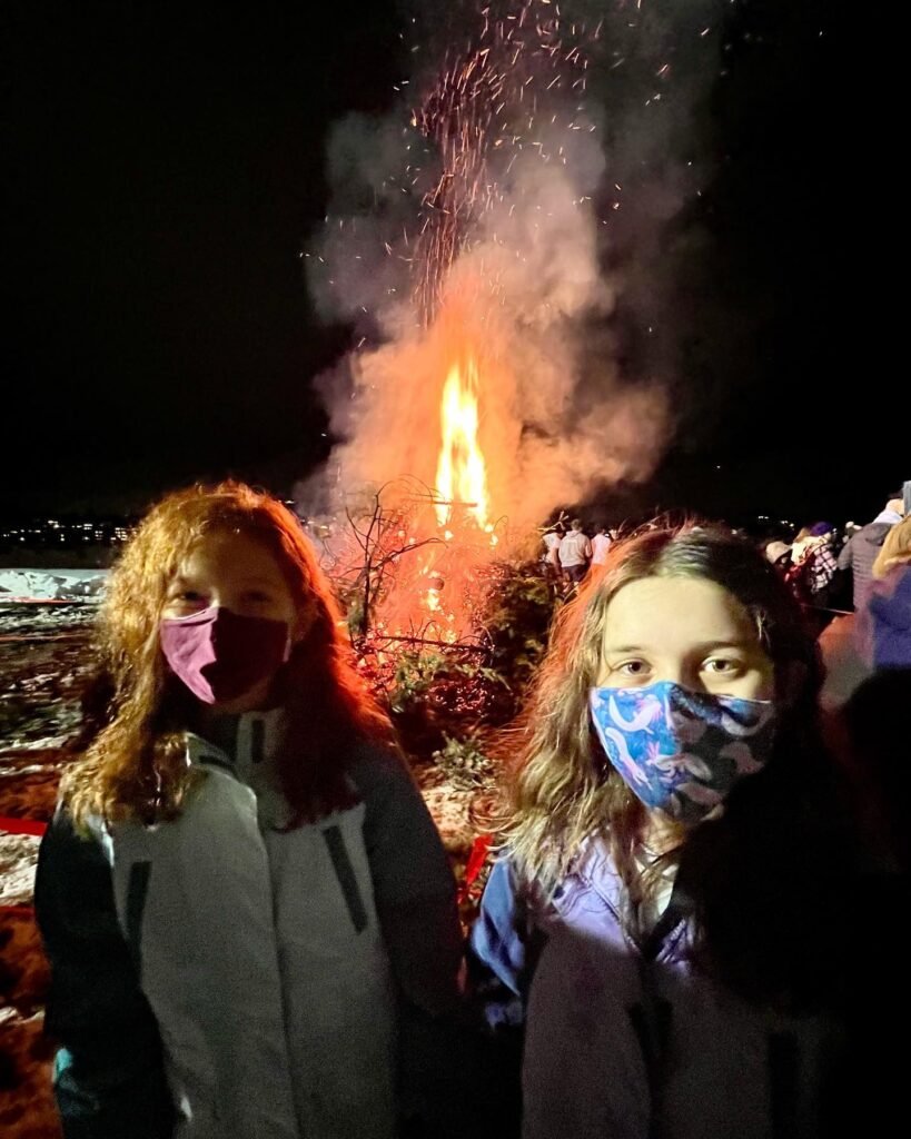 Two children wearing face masks stand in front of a large fire at night.