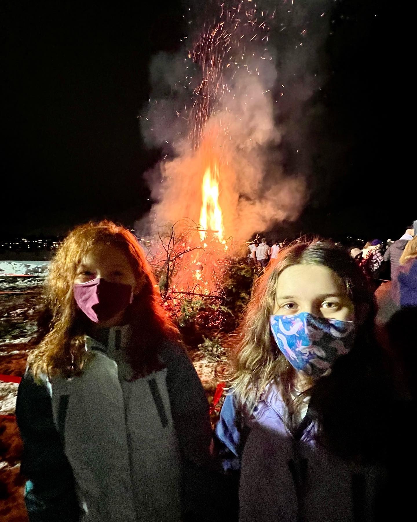 Two children wearing face masks stand in front of a large fire at night.