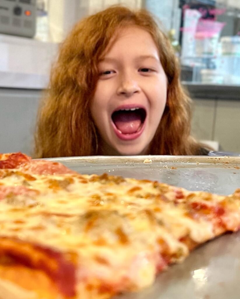 A happy child with red hair smiles broadly behind a tray of homemade cheese pizza.