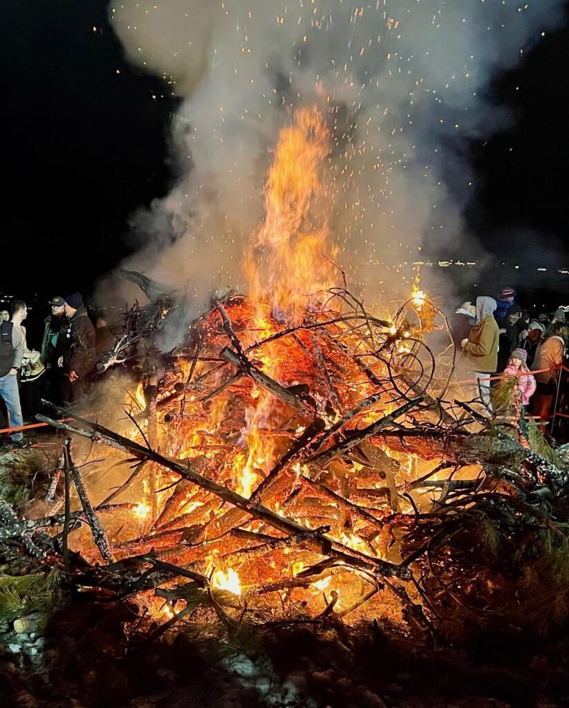 A large nighttime bonfire at a natural site in the Pacific Northwest, with people gathered around the clearing. Smoke and sparks rise into the dark sky in this travel and lifestyle photo.