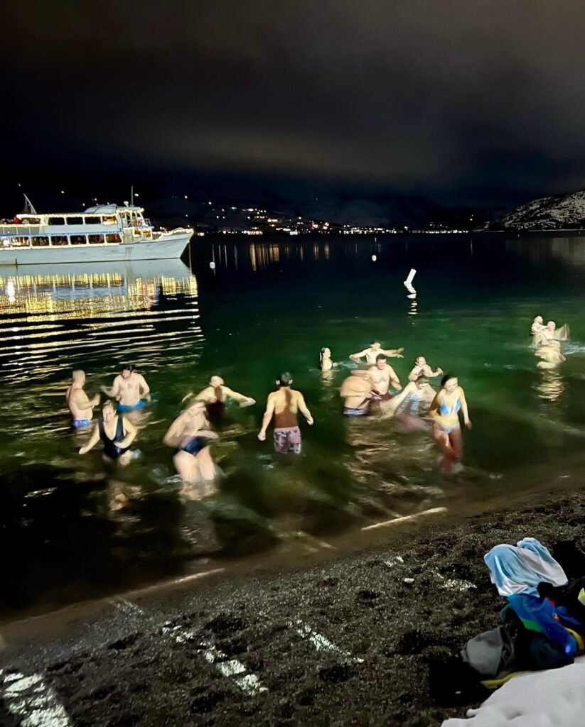 Friends enjoy a nighttime dip from a pebbly shore, illuminated by a nearby boat and shoreline lights, set against a dark, moody bay sky.