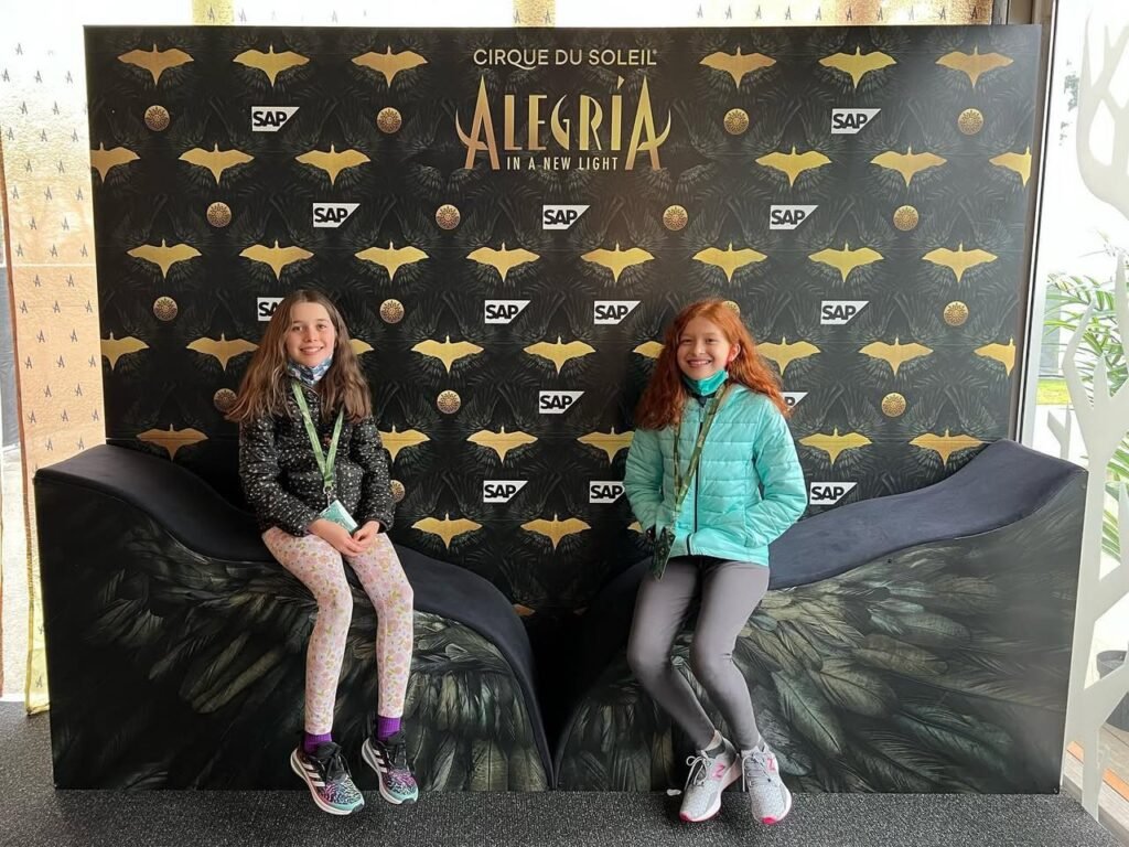 Two people sitting in front of the Cirque Du Soleil Alegria backdrop at a backstage event, wearing casual jackets and leggings.