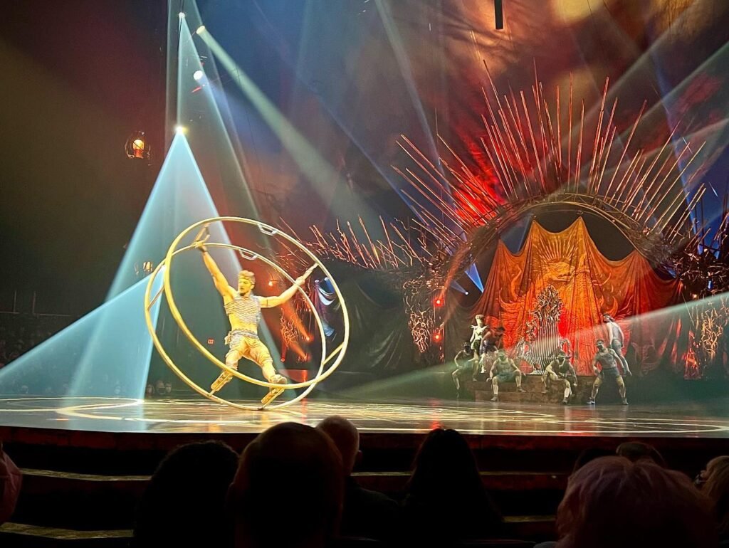 A performer in a golden costume stands on a stage illuminated by dramatic beams of light. Silhouettes of audience members watch the spectacle unfold.