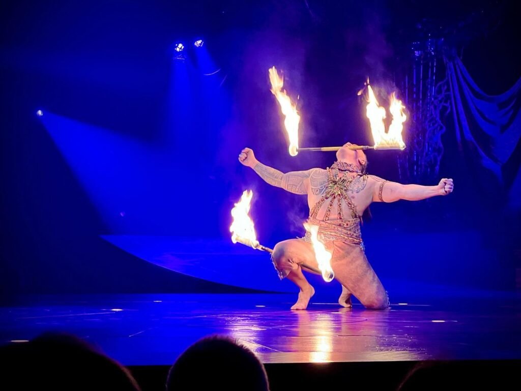 A performer twirling flaming torches on a stage illuminated with blue lights.