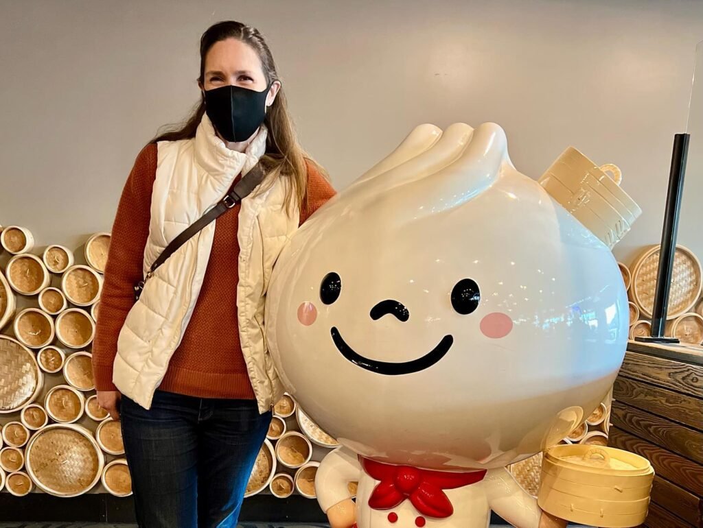 A masked person poses joyfully next to a giant inflatable dim sum character with bamboo steamers visible in the background, highlighting a playful dining atmosphere.