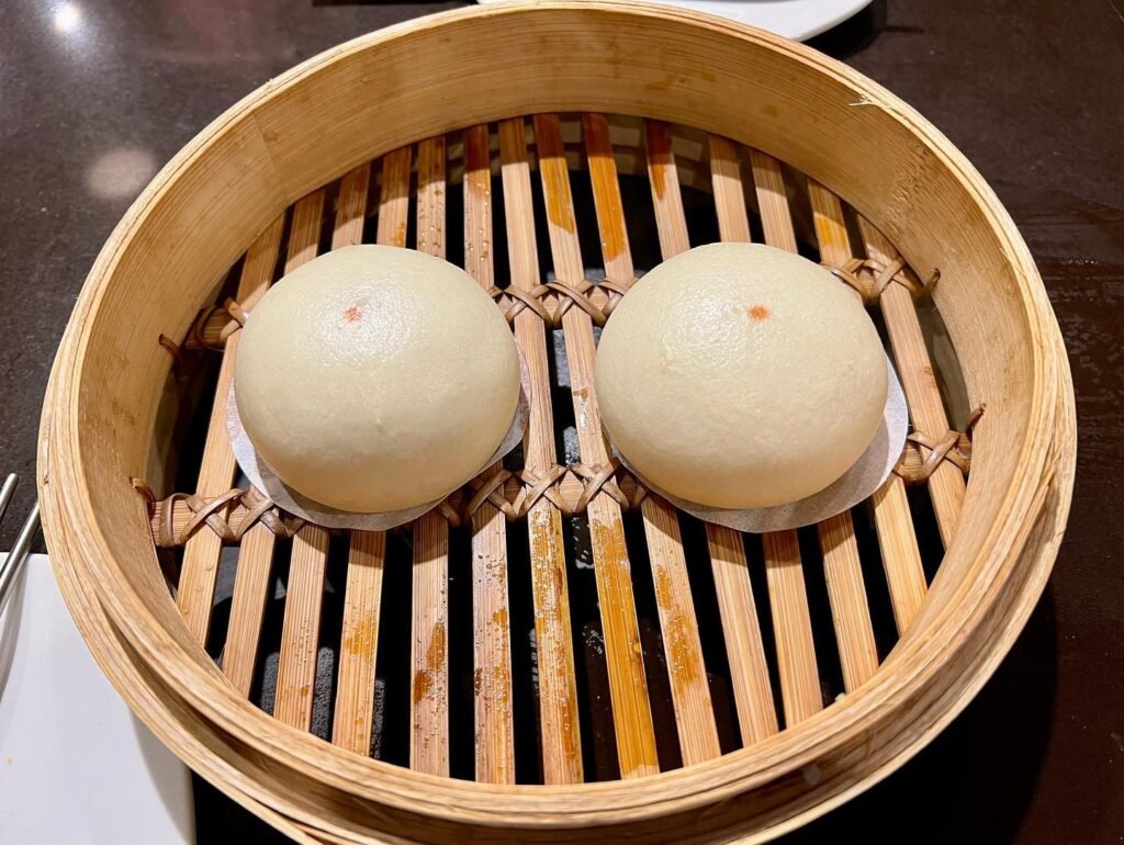 Two round steamed buns sit in a bamboo steamer basket. Close-up view highlights the texture of the dough and bamboo slats.