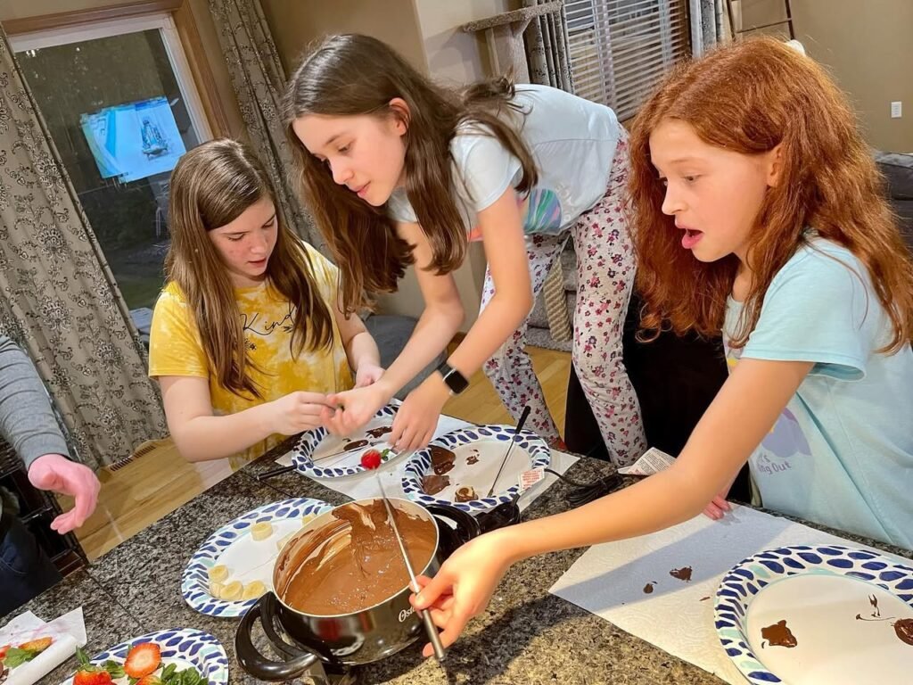 Several children enjoying a hot chocolate fountain at a kitchen island. Dipped strawberries and plates are visible on the counter.