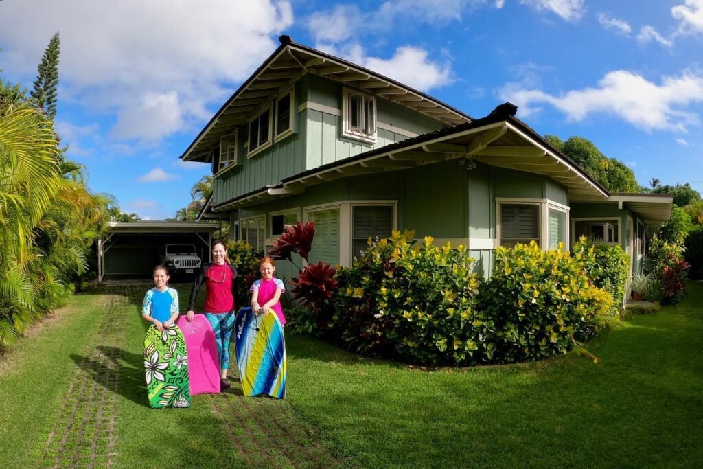Several colorful, hand-painted cloths are being displayed on a lawn in front of a two-story house with a lush garden under a bright blue sky.