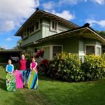 Several colorful, hand-painted cloths are being displayed on a lawn in front of a two-story house with a lush garden under a bright blue sky.