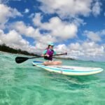 A wide-angle shot of a person on a paddleboard in clear turquoise water under a blue sky, with a second person paddling nearby.