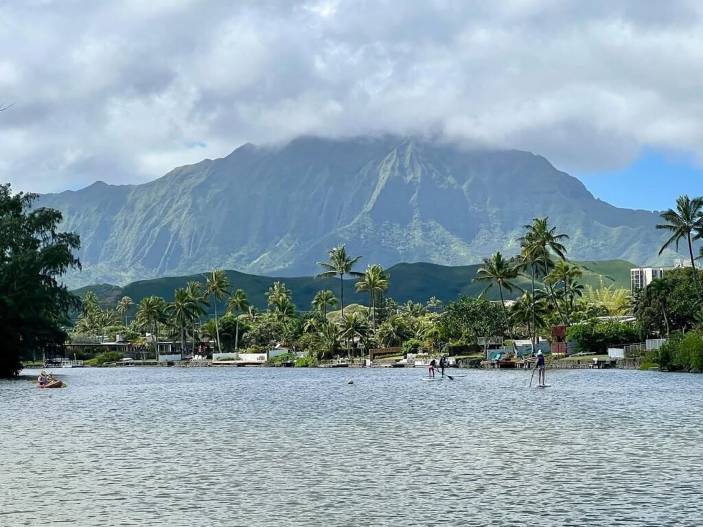 A panoramic view of Diamond Head mountain towering over the water of Oahu Bay, framed by palm trees and a cloudy sky. Several swimmers enjoy the calm lake in the foreground.