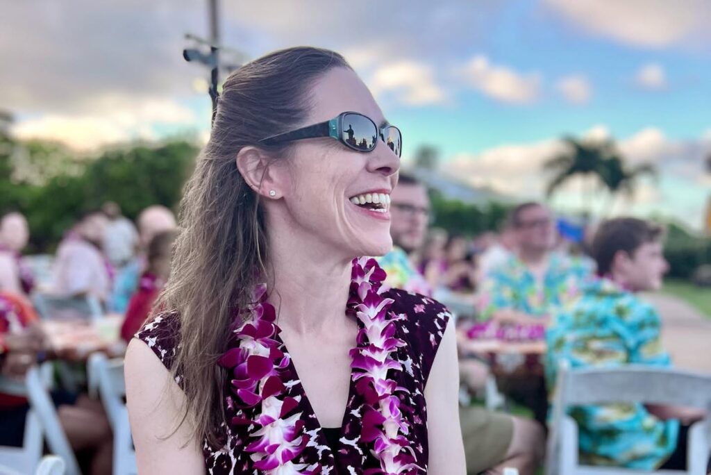 A smiling person wearing sunglasses and a vibrant floral shirt at an outdoor event with palm trees and other guests in the background.