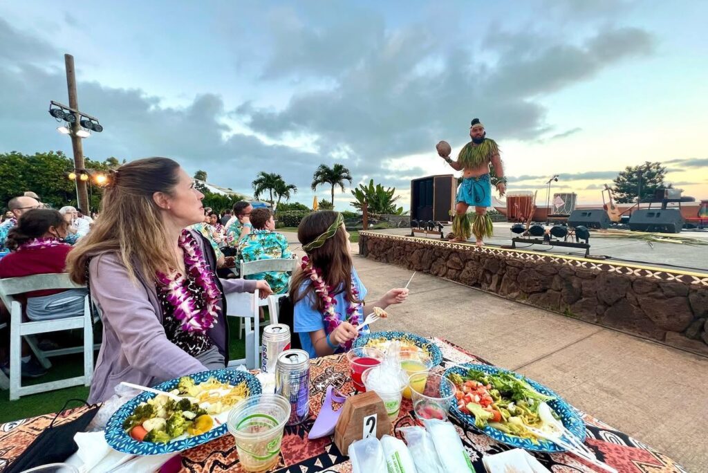 A group dines on colorful platters outdoors as a performer hosts nearby on a stone ledge under a dusk sky. Palm trees and event lighting complete the tropical outdoor dining atmosphere.