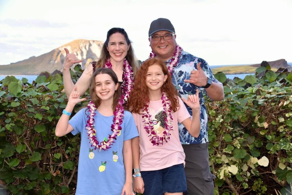 A group of people wearing floral leis posing outdoors with a mountain and lush greenery in the background.