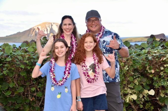 A group of people wearing floral leis posing outdoors with a mountain and lush greenery in the background.