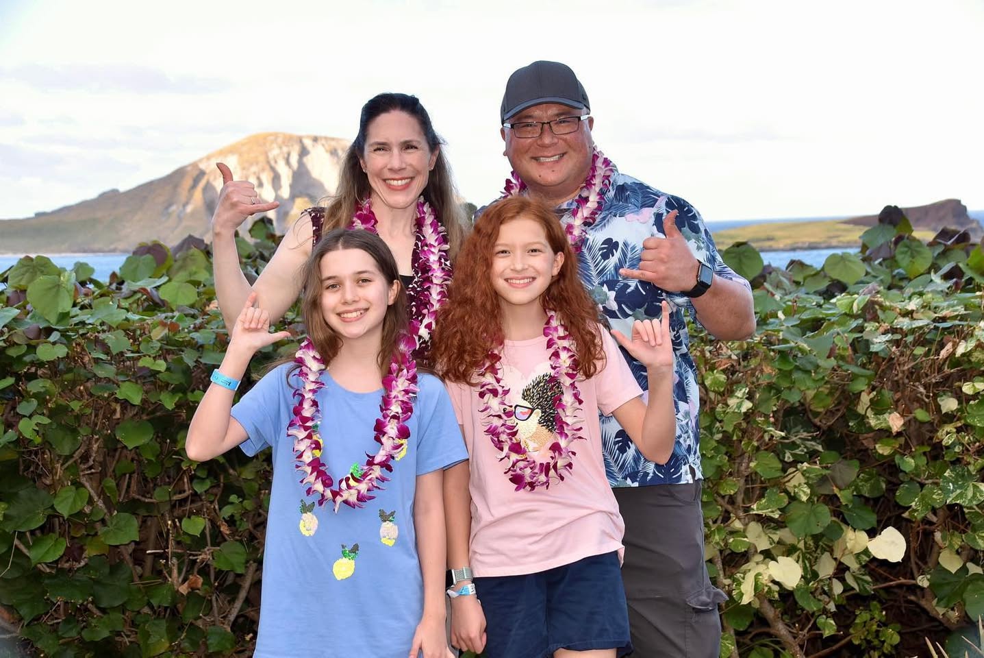 A group of people wearing floral leis posing outdoors with a mountain and lush greenery in the background.