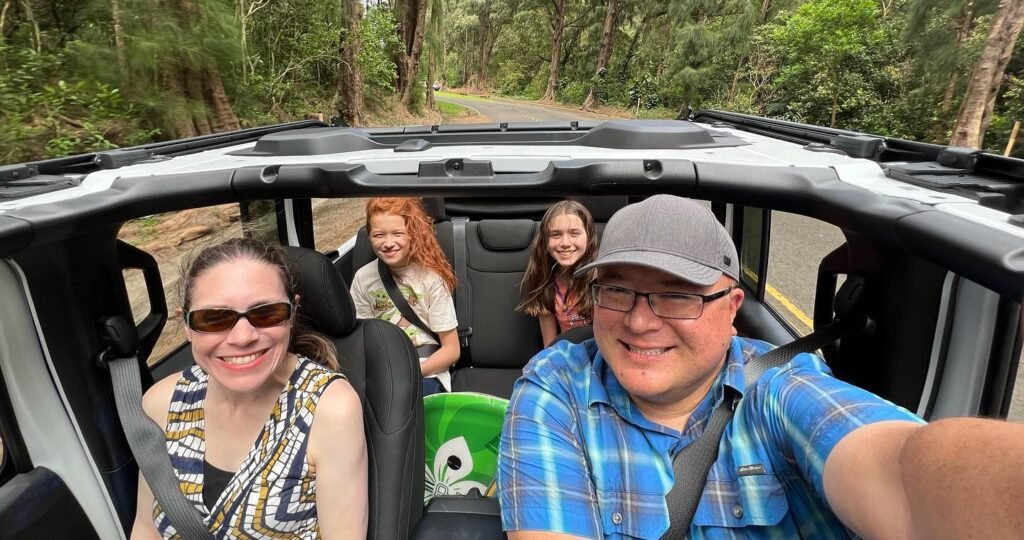 A group of people taking a selfie inside a Jeep during a scenic safari tour. The vehicle is cruising along a dirt road surrounded by dense forest foliage.