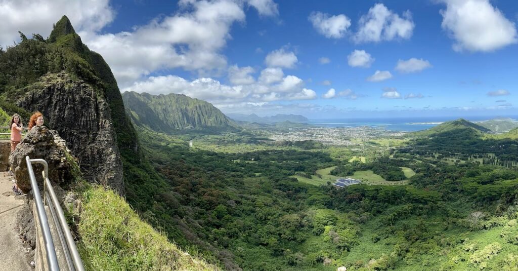 A stunning scenic overlook showing lush green cliffs and valleys leading to the ocean in Hawaii. Visitors enjoy the breathtaking coastal view from an elevated hiking path.