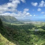 A stunning scenic overlook showing lush green cliffs and valleys leading to the ocean in Hawaii. Visitors enjoy the breathtaking coastal view from an elevated hiking path.