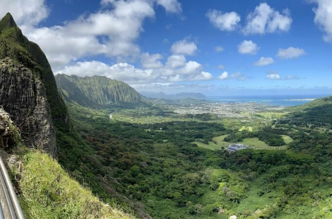 A stunning scenic overlook showing lush green cliffs and valleys leading to the ocean in Hawaii. Visitors enjoy the breathtaking coastal view from an elevated hiking path.