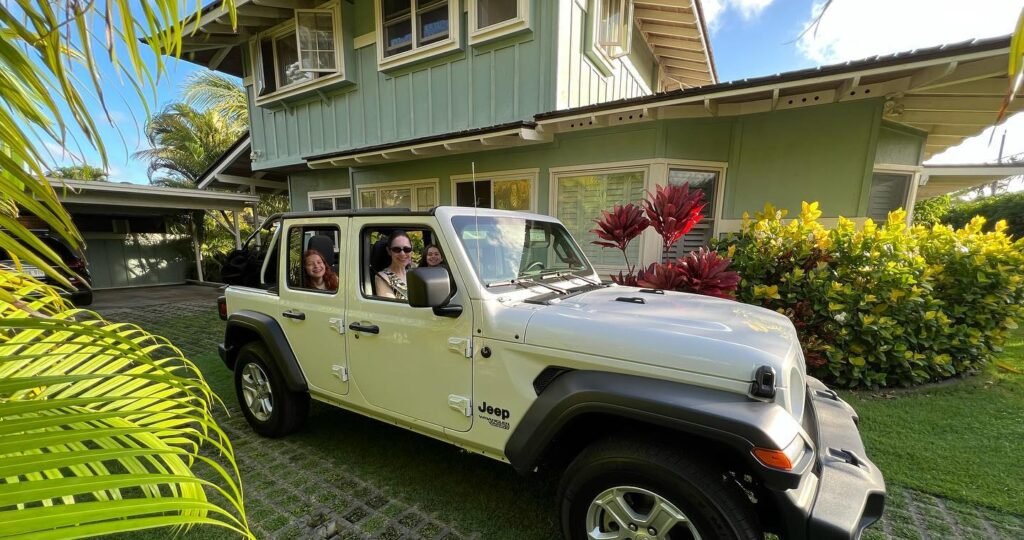 A white Jeep Wrangler parked outside a green house surrounded by lush tropical plants and flowering shrubs under a sunny sky.