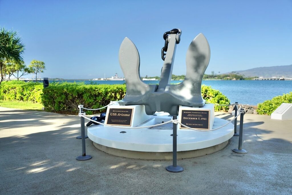 A large anchor monument stands on a paved walkway beside a waterfront park, with a clear blue sky and distant hills in the background.