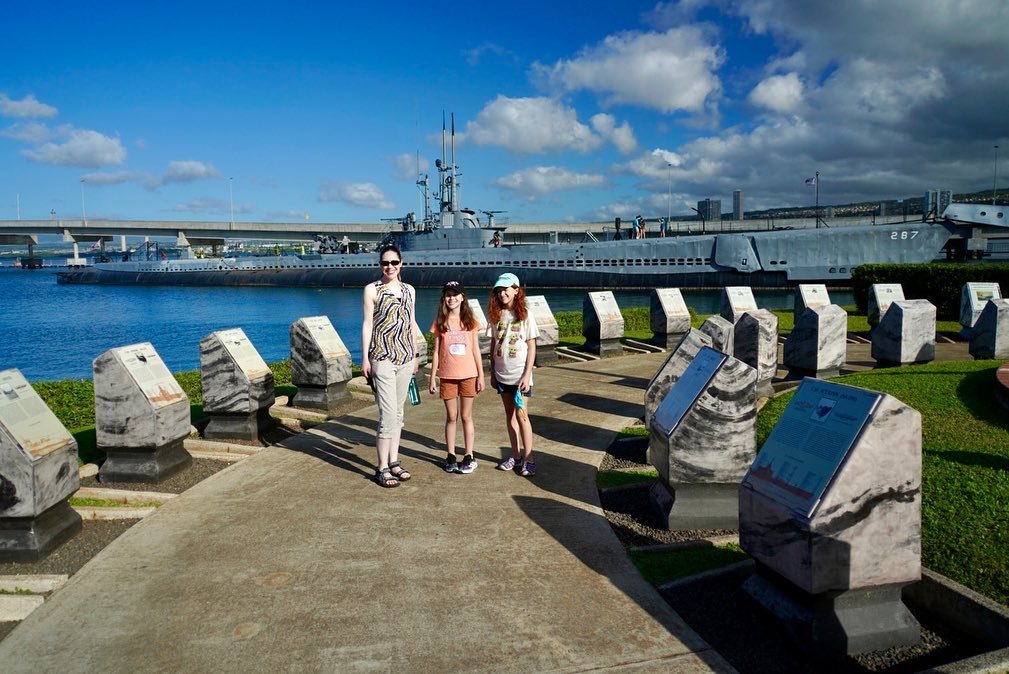 A family walks past stone markers with a Navy ship in the background. A scenic view showcases the memorial's design and waterfront setting.