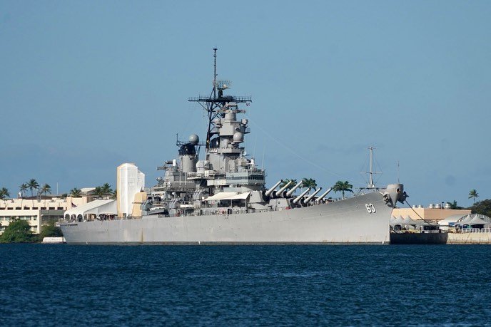 A historic U.S. Navy battleship rests peacefully in tropical waters, framed by palm trees and a clear sky, capturing a moment of maritime history.