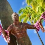 A bronze surfer statue adorned with a pink floral lei stands against a backdrop of palm trees and a clear blue sky. The image captures iconic Hawaiian coastal scenery.