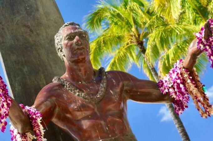 A bronze surfer statue adorned with a pink floral lei stands against a backdrop of palm trees and a clear blue sky. The image captures iconic Hawaiian coastal scenery.
