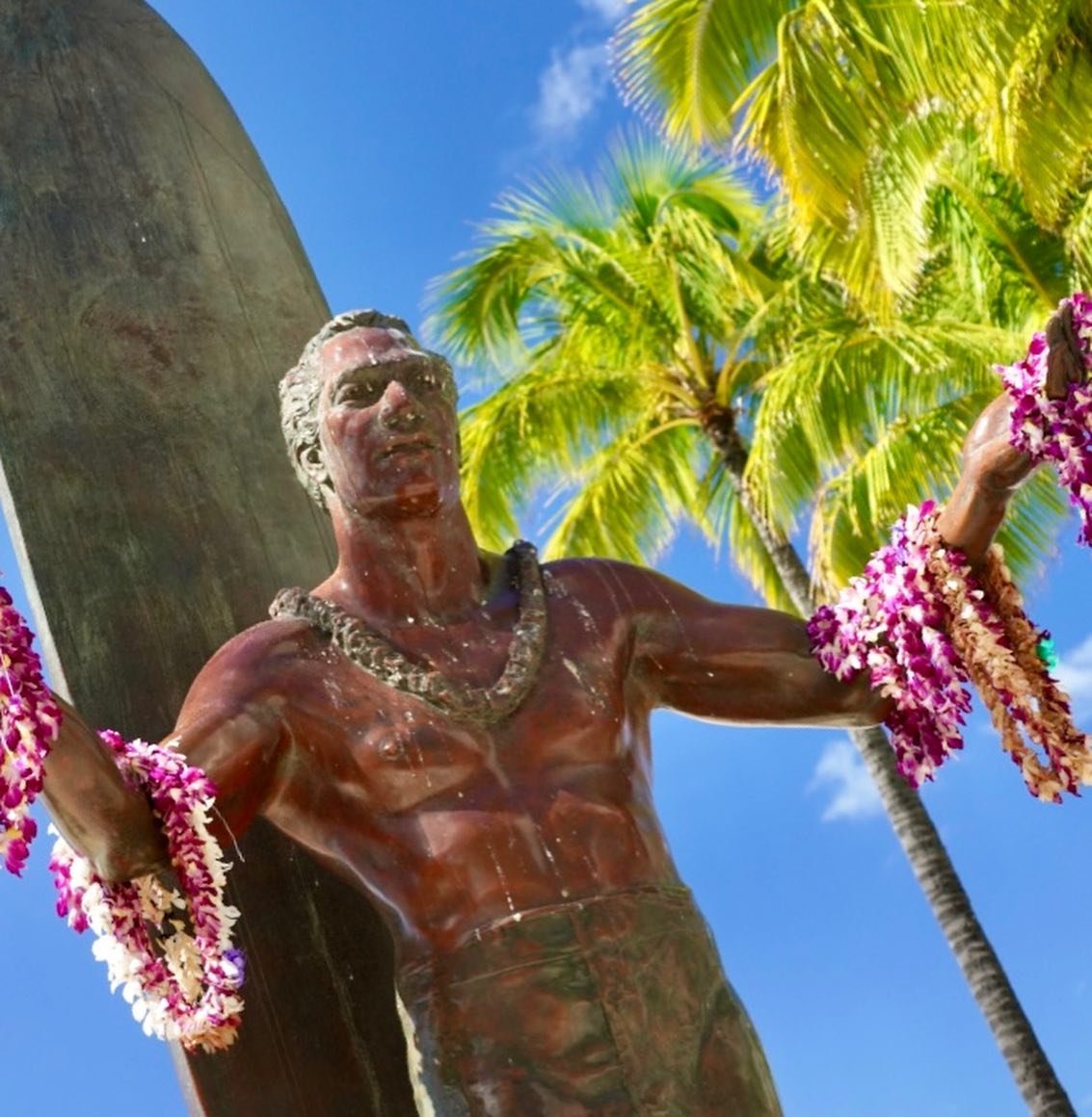 Waikiki A bronze surfer statue adorned with a pink floral lei stands against a backdrop of palm trees and a clear blue sky. The image captures iconic Hawaiian coastal scenery.