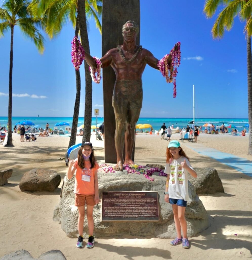 Bronze statue of Tiki adorned with lei on Waikiki Beach with palm trees and turquoise water.